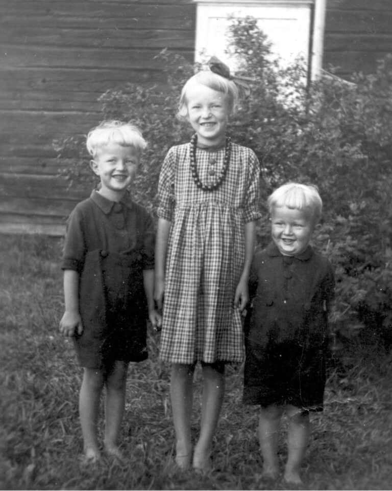 Vanha mustavalkoinen valokuva kolmesta lapsesta, jotka seisovat ulkona ruohikolla puutalon edessä. Keskellä on tyttö ruudullisessa mekossa ja helmikaulakorussa, hänen molemmin puolin seisovat kaksi hymyilevää poikaa tummissa paidoissa. Kaikilla on paljaat jalat, ja he katsovat kameraan iloisina.