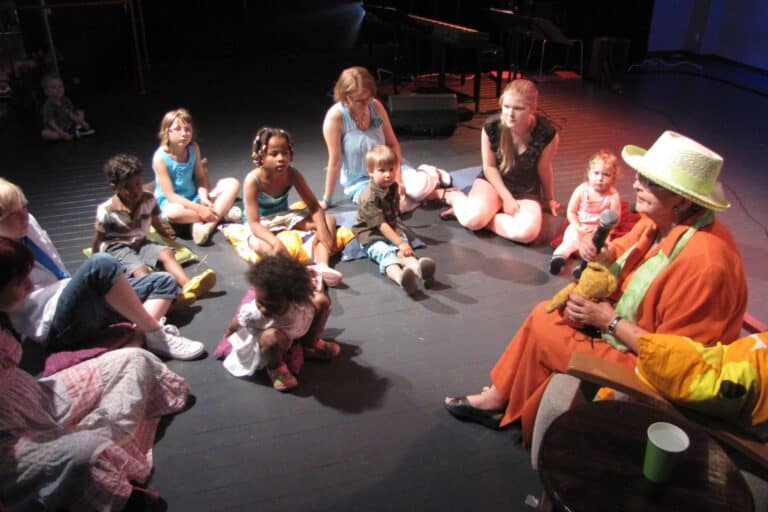 Värivalokuva, jossa iäkäs nainen värikkäissä vaatteissa ja leveälierinen hattu päässään kertoo tarinaa lapsille. Hän istuu tuolissa ja pitelee käsinukkea, ja ympärillä lattialla istuu joukko eri-ikäisiä lapsia ja muutama aikuinen kuuntelemassa keskittyneesti. Taustalla näkyy piano ja hämärä esiintymislava.