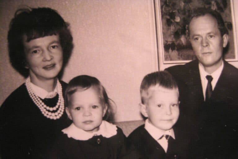 Vanha mustavalkoinen perhevalokuva, jossa nelihenkinen perhe istuu yhdessä. Vasemmalla on äiti helmikaulakorussa ja tummassa puvussa, hänen vieressään pieni tyttö valkokauluksisessa mekossa. Oikealla puolella istuvat poika ja isä, molemmat pukeutuneina tummaan pukuun ja valkoiseen paitaan. Taustalla näkyy taulu seinällä.