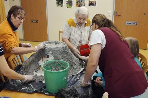 Ryhmä iäkkäitä ihmisiä ja ohjaaja työskentelevät yhdessä pöydän ääressä tehden suurta harmaata massarakennelmaa, joka muistuttaa vuorta. Pöydällä on musta suojamuovi ja vihreä ämpäri, ja osallistujat käyttävät suojakäsineitä. Kuva on otettu sisätilassa, ja tunnelma on keskittynyt ja yhteisöllinen.