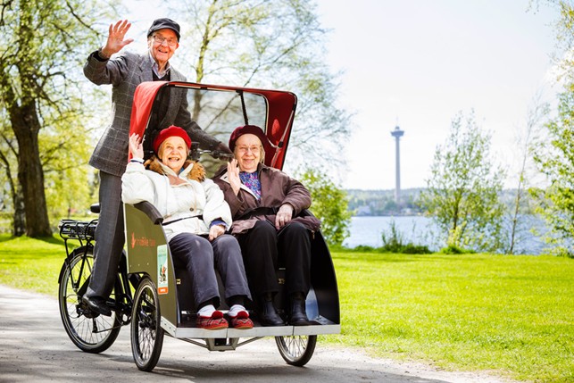 Kaksi iäkästä ihmistä istuu pyöräriksan kyydissä ja hymyilee iloisesti, kun taustalla näkyy järvimaisema ja vehreä puisto. Pyöräriksaa ohjaa kolmas henkilö, joka vilkuttaa kameralle. Kuvan taustalla erottuu Tampereen Näsinneula-torni. Tunnelma on aurinkoinen ja hyväntuulinen.