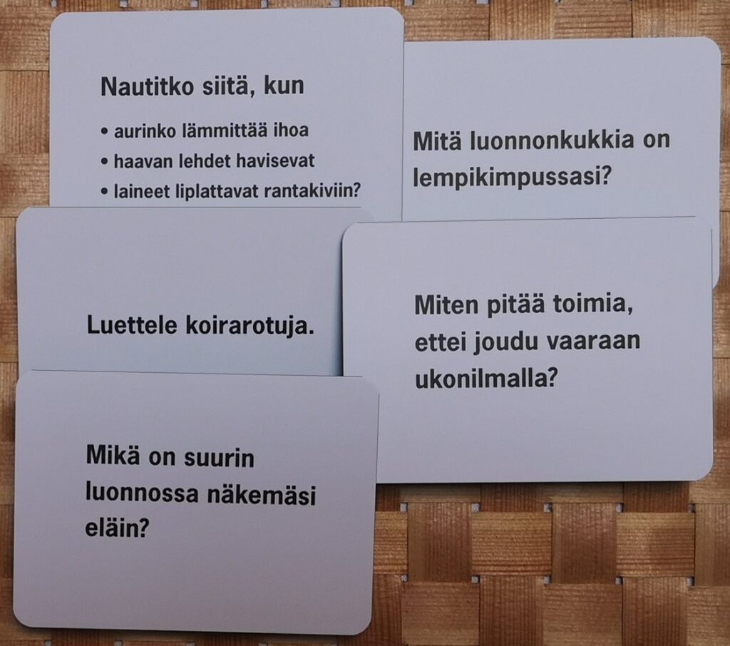 Vaaleanharmaita keskustelukortteja, joissa on mustalla painettuja kysymyksiä luontoon ja arkeen liittyen. Tekstit näkyvät selkeästi: “Nautitko siitä, kun aurinko lämmittää ihoa, haavan lehdet havisevat, laineet liplattavat rantakiviin?”, “Mitä luonnonkukkia on lempikimpussasi?”, “Luettele koirarotuja.”, “Miten pitää toimia, ettei joudu vaaraan ukonilmalla?” ja “Mikä on suurin luonnossa näkemäsi eläin?”. Kortit ovat päällekkäin korimaisen taustan päällä.