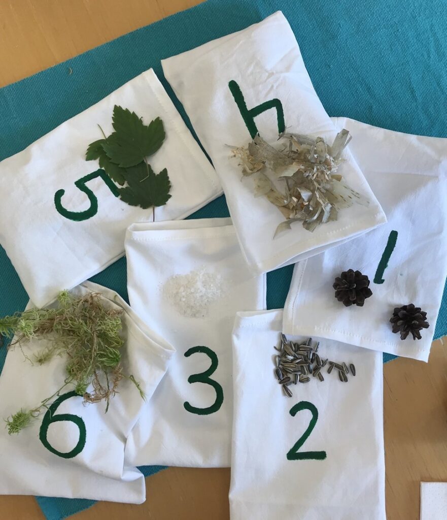 Valkoisia kangaspusseja, joiden päälle on maalattu vihreillä numeroilla luvut 1–6. Jokaisen pussin päällä on jokin luonnosta kerätty esine tunnusteltavaksi: numerolla 1 pieniä käpyjä, numerolla 2 auringonkukansiemeniä, numerolla 3 karkeaa suolaa, numerolla 5 vihreitä lehtiä ja numerolla 6 sammalta. Pussit ovat aseteltu turkoosin kankaan päälle pöydälle.